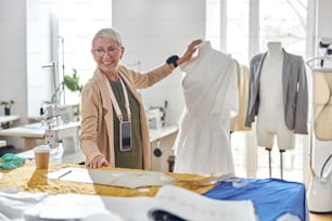Smiling short haired mature fashion designer with elegant glasses stands between mannequin with white fabric and cutting table in spacious sewing workshop