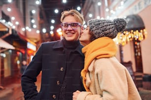 Young girl standing on christmas decorated street and kisses her boyfriend.
