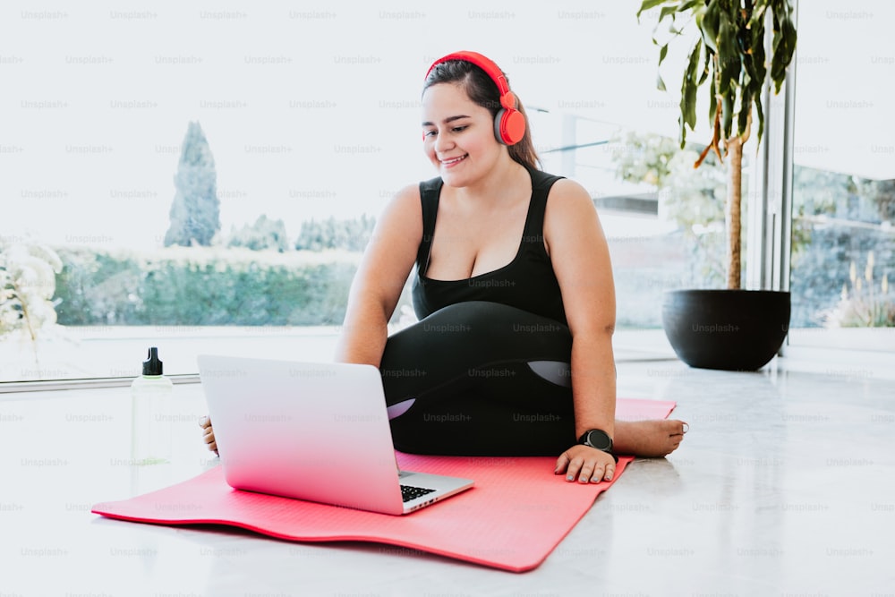 Ragazza formosa ispanica con cuffie rosse e abbigliamento sportivo che prende lezioni di yoga online sul laptop a casa