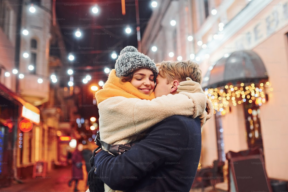 Positives junges Paar in warmen Kleidern, das sich auf der weihnachtlich geschmückten Straße umarmt.