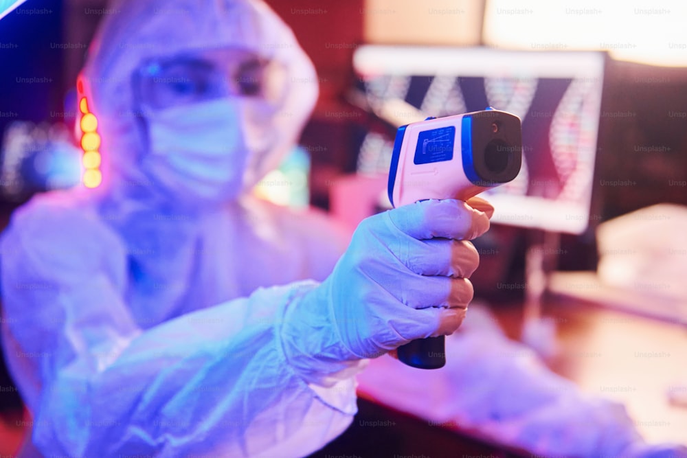 Enfermeira de máscara e uniforme branco, segurando termômetro infravermelho e sentada em laboratório iluminado por neon com informática e equipamentos médicos em busca da vacina contra o Coronavírus.