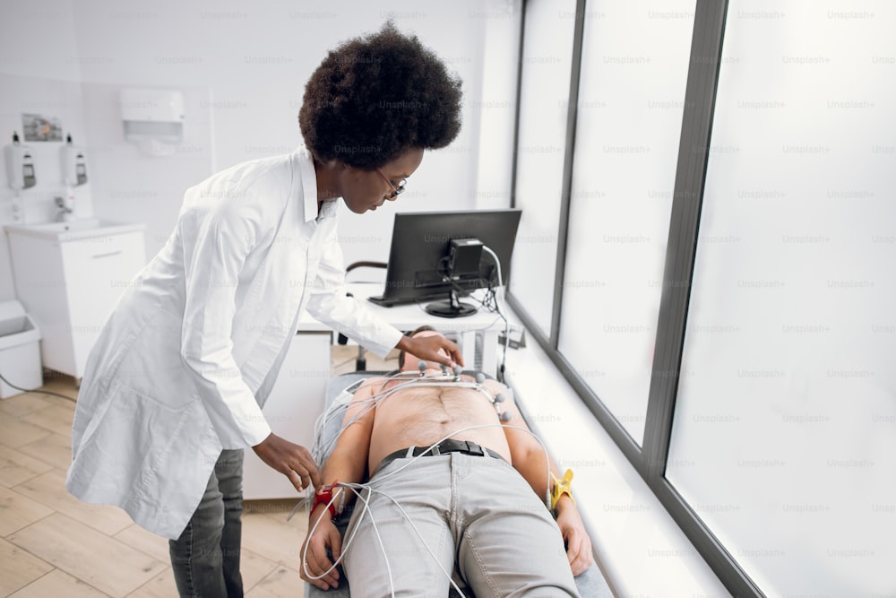 현대 클리닉에서 청년의 ECG 테스트. 젊은 아프리카 여자 의사 심장 전문의는 ECG를 만들기 전에 남성 백인 환자의 가슴에 특수 장비, 진공 센서 전극을 넣습니다.