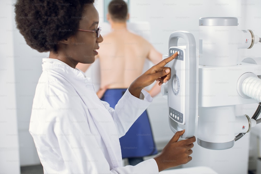 젊은 아프리카계 미국인 여성 의사 방사선 전문의의 측면 보기를 닫고 현대 엑스레이 기계의 제어판에 있는 버튼을 누릅니다. 배경에 남성 환자의 흐릿한 보기
