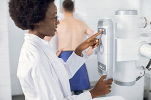 Vue latérale rapprochée d’une jeune femme afro-américaine médecin radiologue, appuyant sur des boutons sur le panneau de commande d’un appareil à rayons X moderne. Vue floue d’un patient masculin à l’arrière-plan