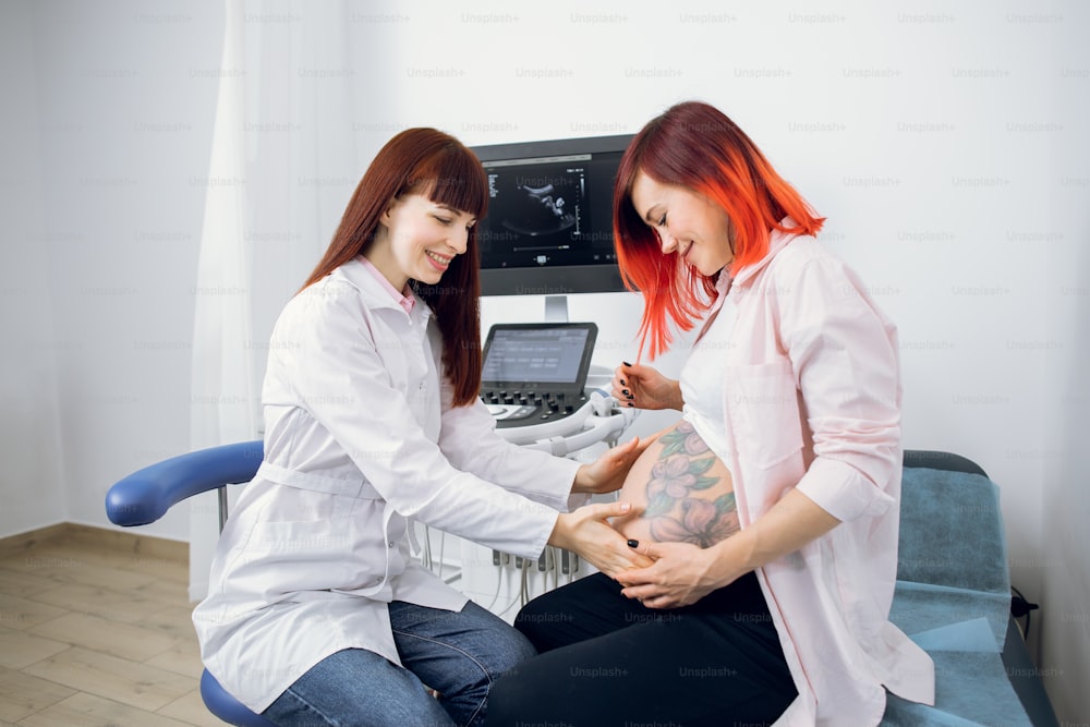 Young professional female Caucasian doctor touches the belly of a pregnant woman during regular check up at modern obstetrics center. Pretty joyful red haired pregnant woman talking with doctor