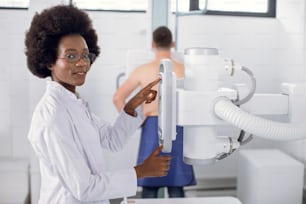 Tecnico radiologo americano Aro femminile che installa una macchina per eseguire la radiografia del torace su un paziente di sesso maschile nella moderna sala di radiologia dell'ospedale