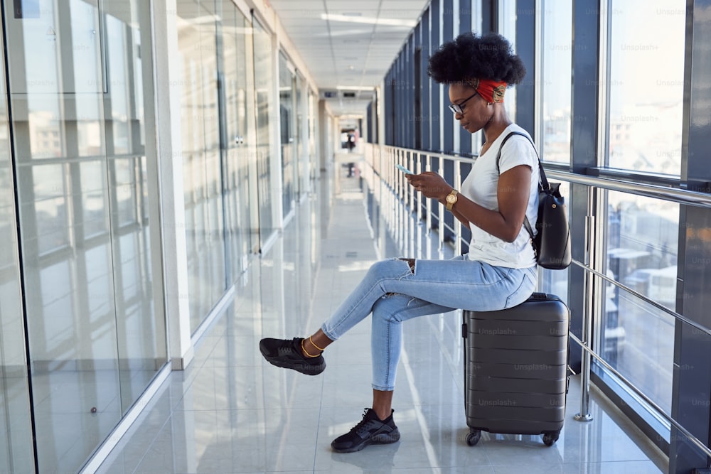 캐주얼 옷을 입은 젊은 아프리카 계 미국인 여성 통행인이 수하물을 가지고 공항에 있습니다. 전화 사용.