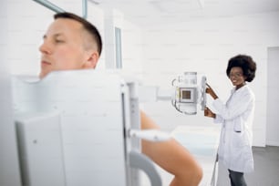 Junger Mann steht im modernen Krankenhaus während des Röntgen-Thorax-Scans, während weibliche afroamerikanische Medizintechnikerin mit Röntgengerät arbeitet. Scannen auf Frakturen, Brust, Krebs oder Tumor