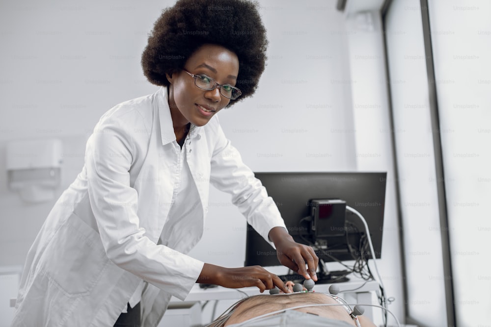 Close up portrait of young pleasant Afro American woman doctor gp cardiologist, sticking ECG electrodes for heart diagnostic for young man patient. Medical equipment for electrocardiogram