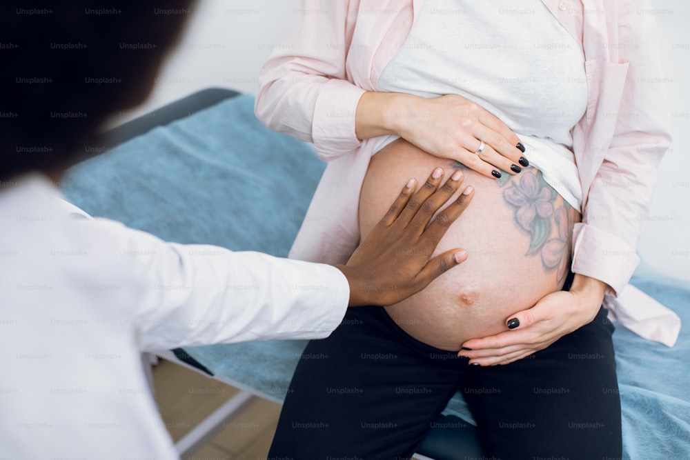 Cropped close up image of unrecognizable female black doctor obstetrician, examining young pregnant woman sitting on couch in hospital. Cute pregnant tummy with flowers tattoo