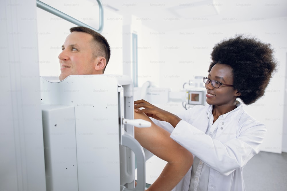 X - ray medical equipment in modern clinic. Female smiling Afro American doctor standing near apparatus, helps male patient during x-ray chest scan. Closeup