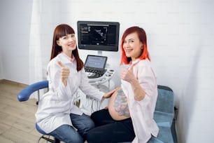 밝은 빨간 머리와 배에 문신을 한 임산부가 임신 검진을 위해 여성 친화적 인 의사를 방문합니다. 여성 조산사와 그녀의 임신한 환자가 웃으며 엄지손가락을 치켜들고 있다