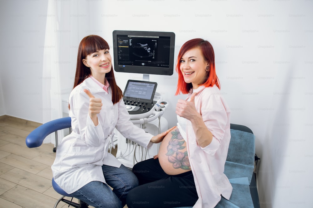 Pregnant lady with bright red hair and tattoo on the belly visiting her female friendly doctor for check up of her pregnancy. Woman midwife and her pregnant patient smiling and showing thumbs up