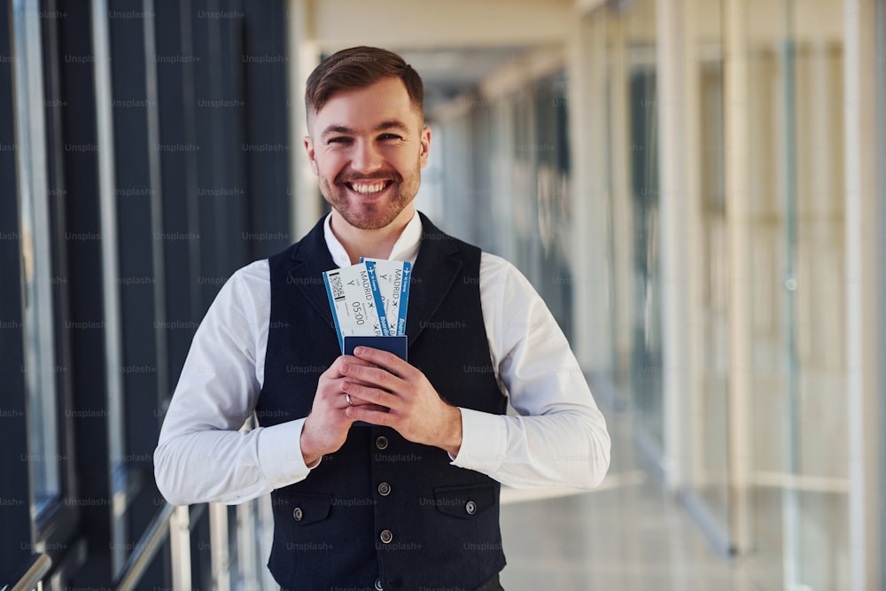 Young male passenger in elegant formal clothes smiling and holding tickets in hands in the airport hall.