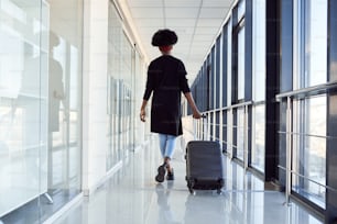 Rear view of young african american female passanger in casual clothes that is in airport with baggage.