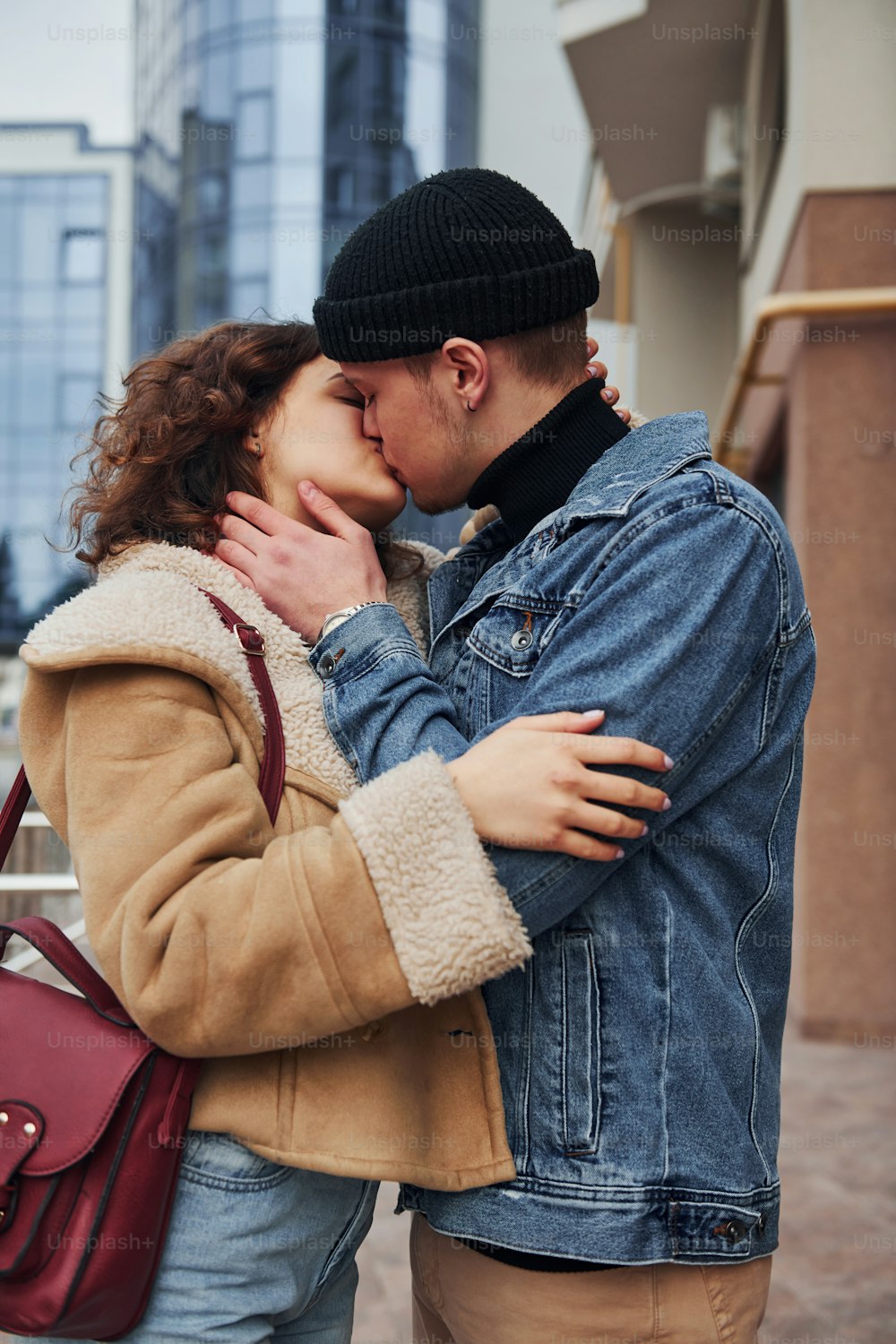 Cheerful couple in casual warm clothes kissing outdoors in the city near business building.