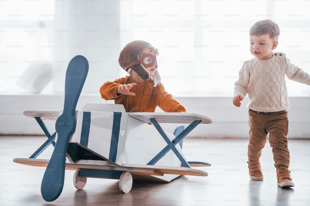 Two little boys in retro pilot uniform having fun with toy plane indoors.