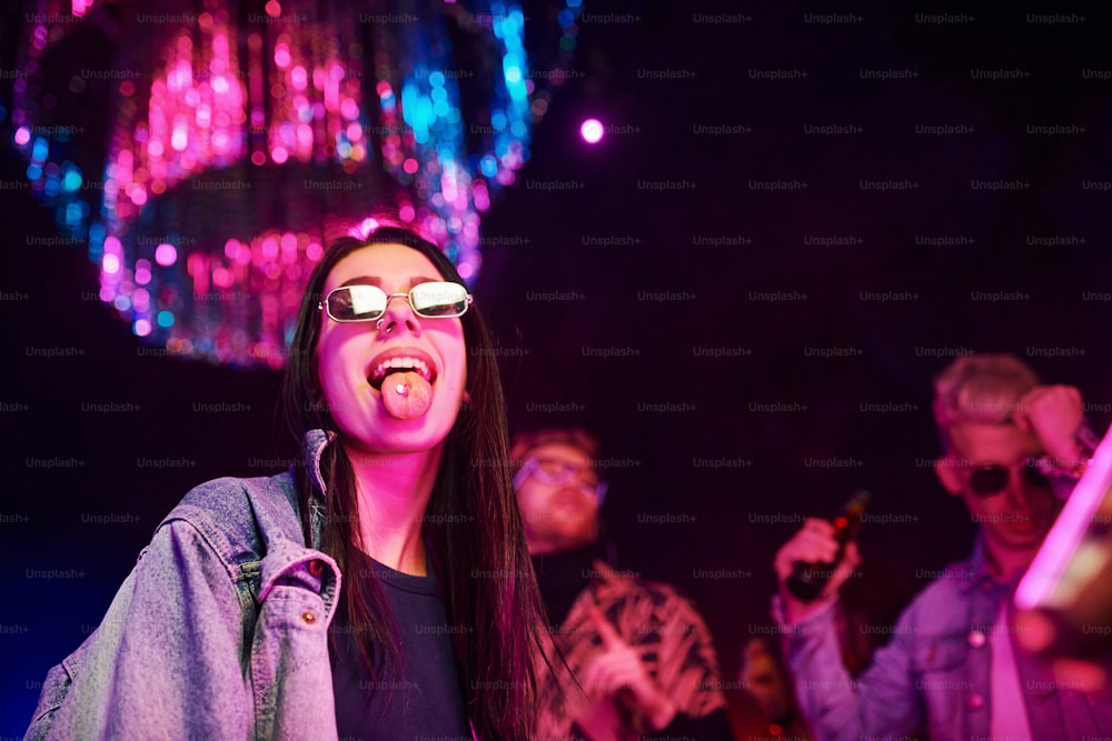 Muchacha joven sosteniendo píldoras de droga en togue frente a jóvenes que se divierten en un club nocturno con luces láser de colores.