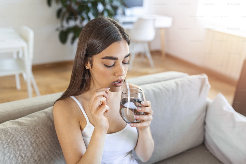 Concept of suffering from terrible pain. Beautiful, sad, upset, unhappy, troubled, weak woman with black hair, wearing casual clothes is sitting on a sofa and taking a pill