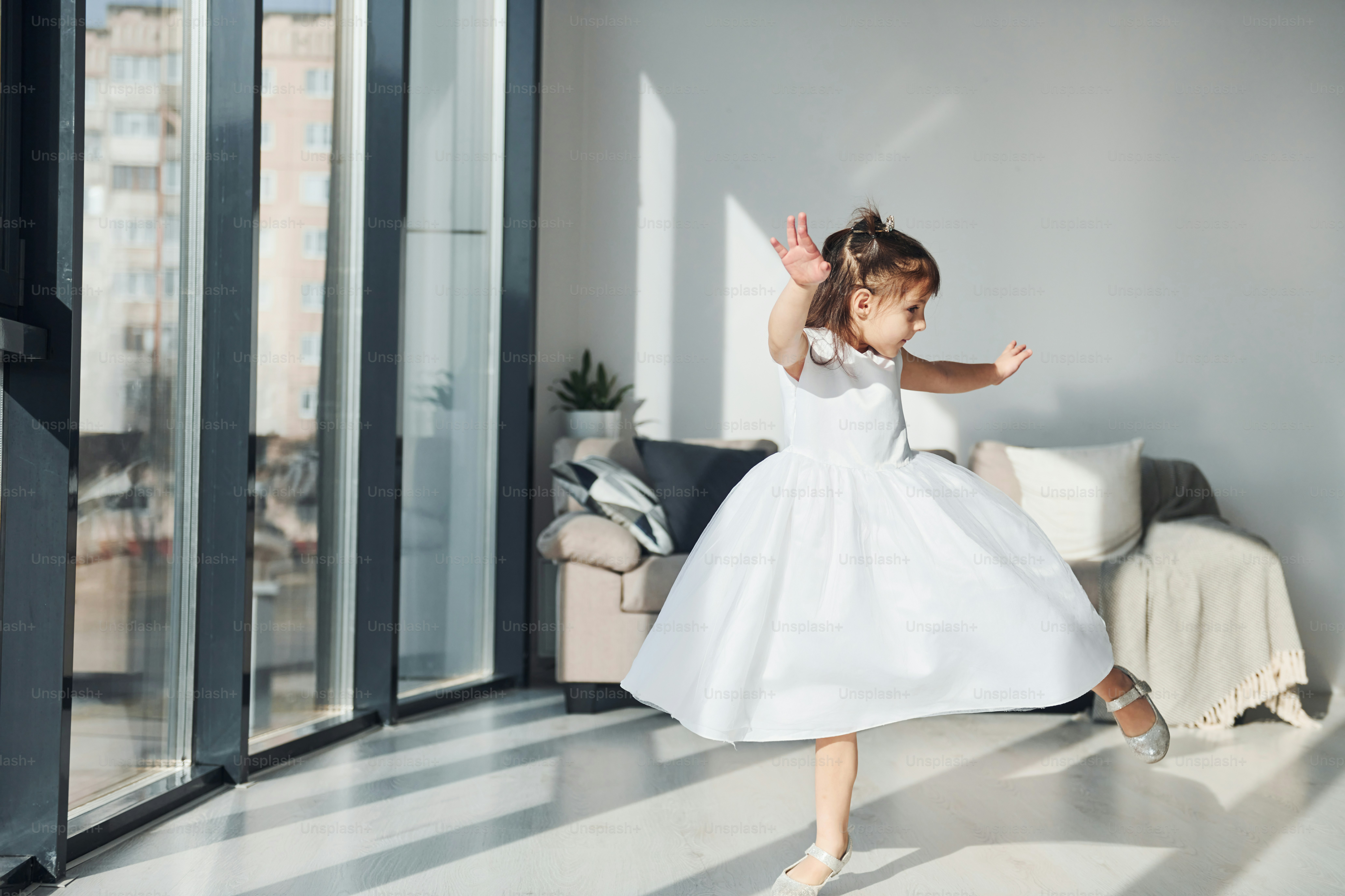 Bambina carina in vestito bianco che balla all'interno in camera da letto da sola.
