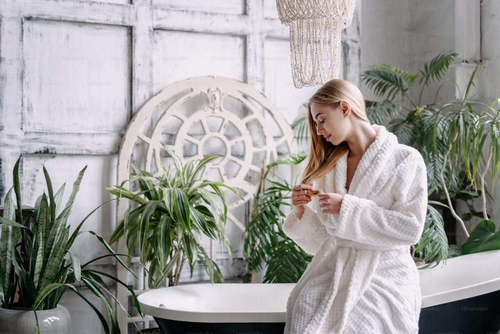 Haircare treatment concept. Calm young woman in white soft bathrobe holding healthy section of hair, checking damage, looking at the cut tips, spending morning in bathroom