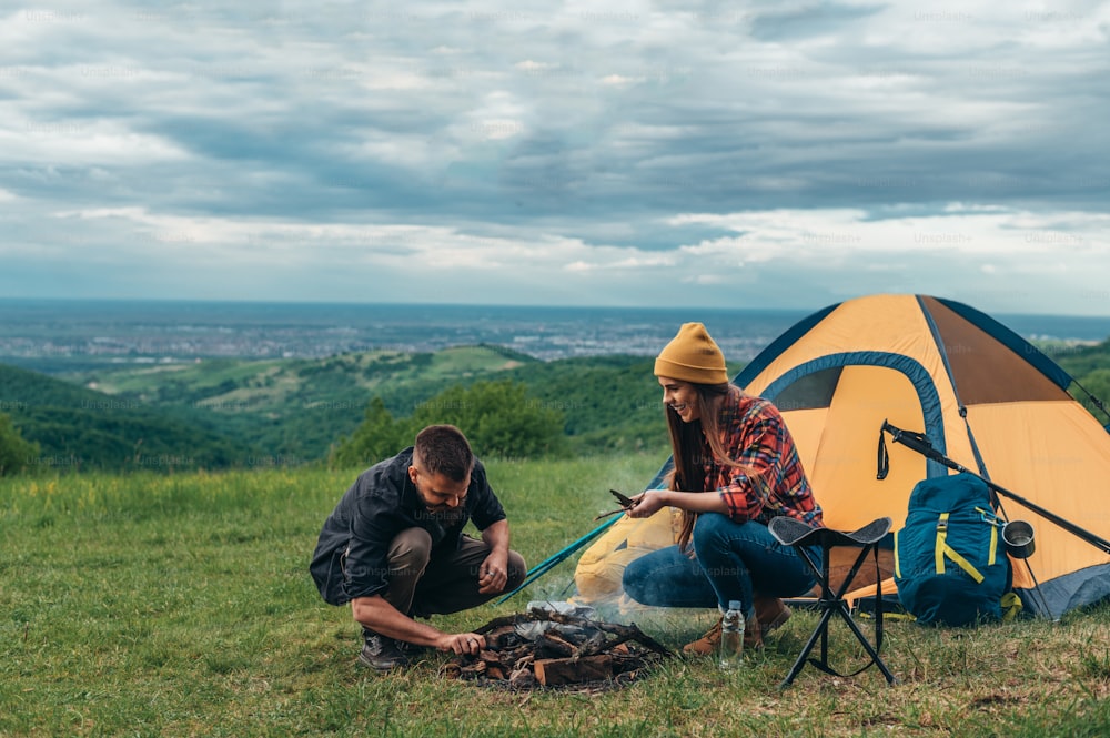 Joven pareja de campistas encendiendo un fuego después de montar su tienda de campaña mientras acampan en la naturaleza