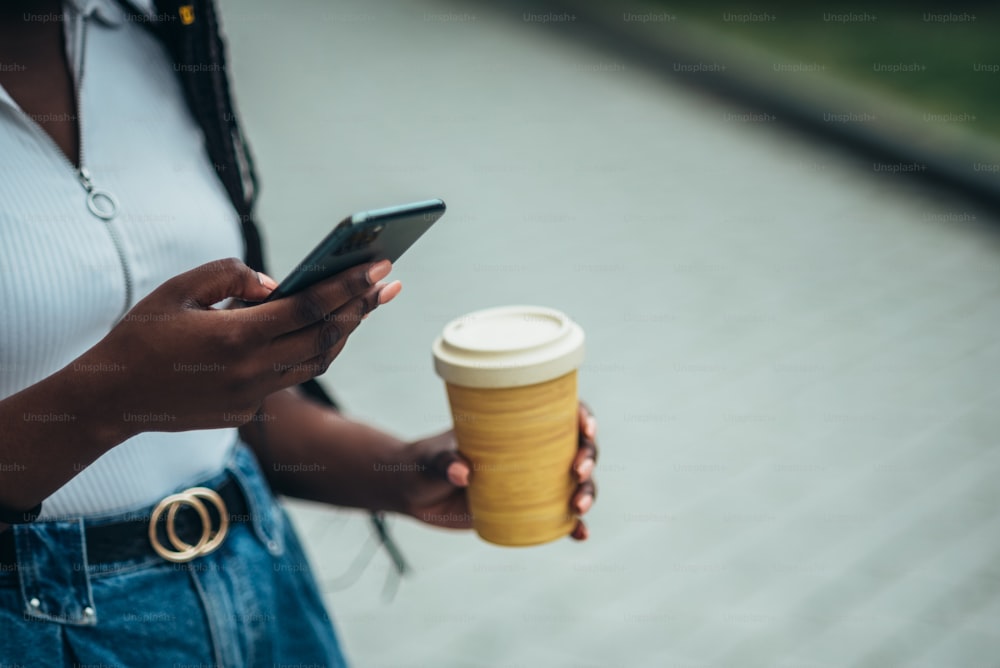 스마트폰을 사용하고 밖에 있는 동안 재사용 가능한 에코 커피 컵을 들고 있는 인식할 수 없는 아프리카계 미국인 여성