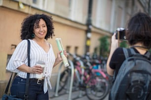 Smiling woman standing on the street with cup of coffee. Woman taking photo of her.