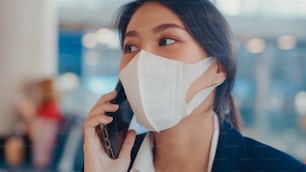 Smart asian business girl wear suit sitting with suitcase in bench call smart phone talk with partner wait for flight at airport. Business travel commuter in covid pandemic, Business travel concept.