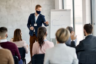 Business leader with face mask giving presentation to group of colleagues while one of them is raising hand to ask him something.