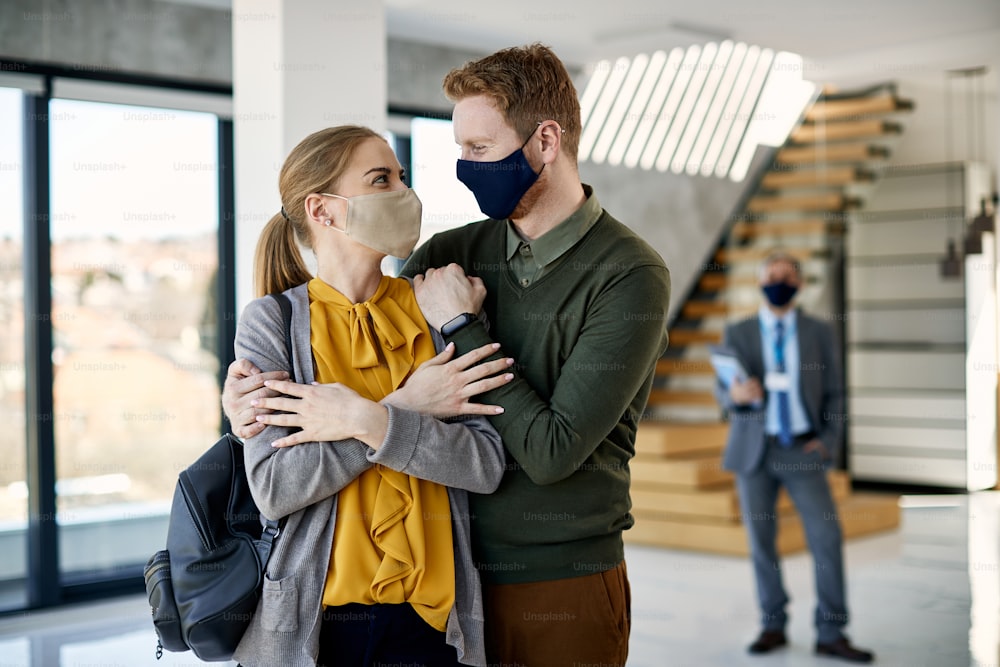 Young embraced couple wearing protective face masks and looking at each other while buying new apartment. Real estate agent is in the background.