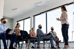 Abaixo veja a líder empresarial com máscara facial se comunicando com o grupo de seus colegas de trabalho em um seminário na sala de conferências.