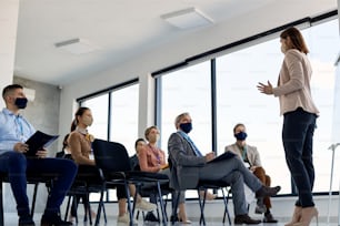 Below view of business leader with face mask communicating with group of her coworkers on a  seminar in conference hall.