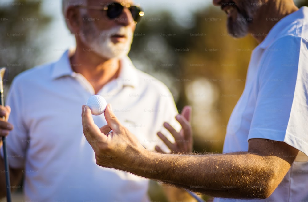 Two older men stand on a golf course and talk. Man holding golf ball. Focus is on foreground.