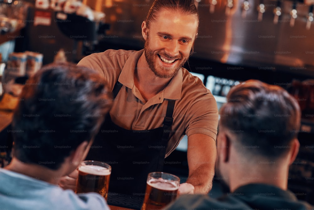 Barman alegre servindo cerveja para homens jovens enquanto está no balcão do bar no pub