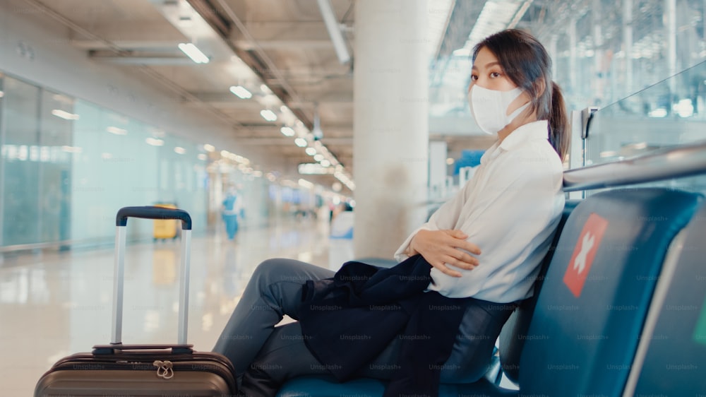 아시아 비즈니스 소녀는 벤치에 앉아 수하물을 들고 걷고 공항에서 비행을 위해 파트너를 찾습니다. 비즈니스 여행 통근 covid 전염병, 비즈니스 여행 사회적 거리두기, 비즈니스 여행 개념.