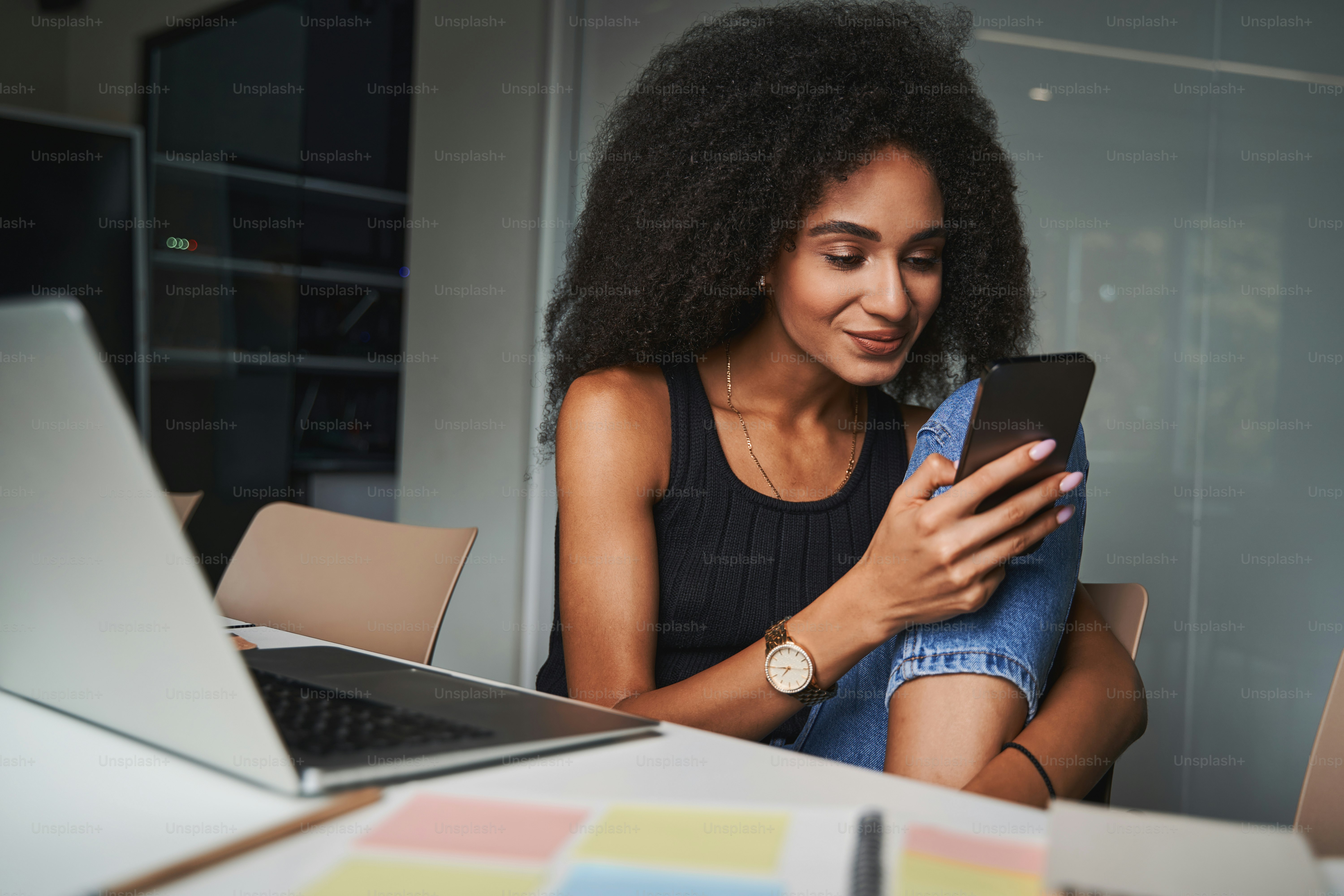 Wanita muda cantik berbalik dari laptop sambil duduk di kantor dan melihat layar smartphone
