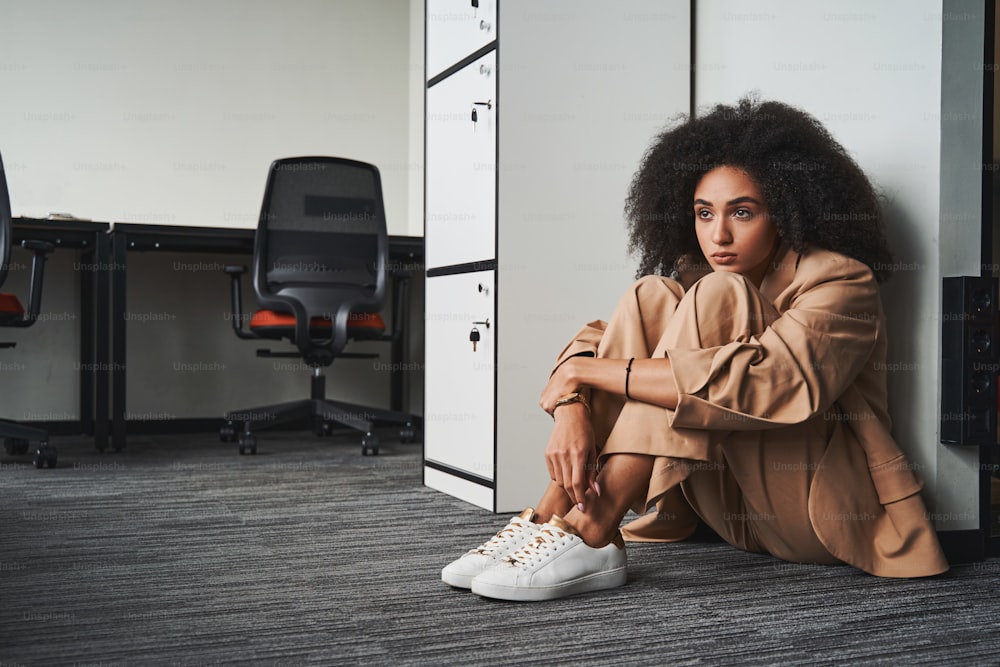 Beautiful woman in a suit sitting on the floor at work and embracing her legs while looking away