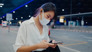 Asian business girl arrive destination wear face mask stand outside look smart phone wait car terminal at domestic airport. Business commuter covid pandemic, Business travel social distancing concept.