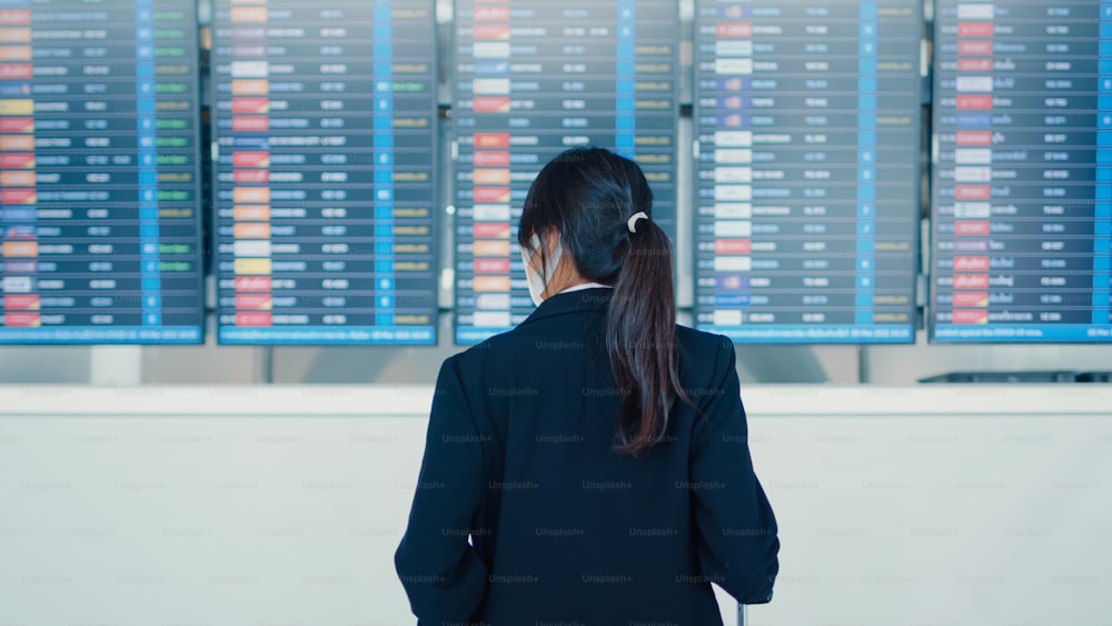 아시아 비즈니스 소녀는 국제 공항에서 그녀의 비행을 확인하는 정보를 보는 보드 앞에 여행 가방 스탠드와 얼굴 마스크를 착용. 비즈니스 통근자 covid 전염병, 비즈니스 여행 개념.