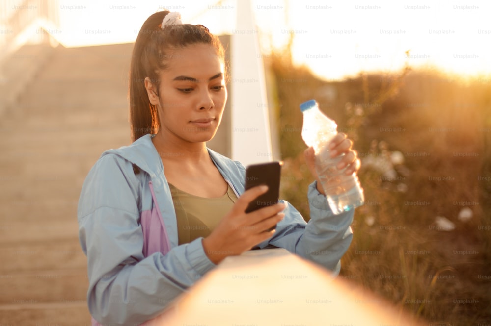 Femme sportive africaine debout sur les escaliers tenant une bouteille d’eau et utilisant un téléphone intelligent. Se tenir au courant des médias sociaux.