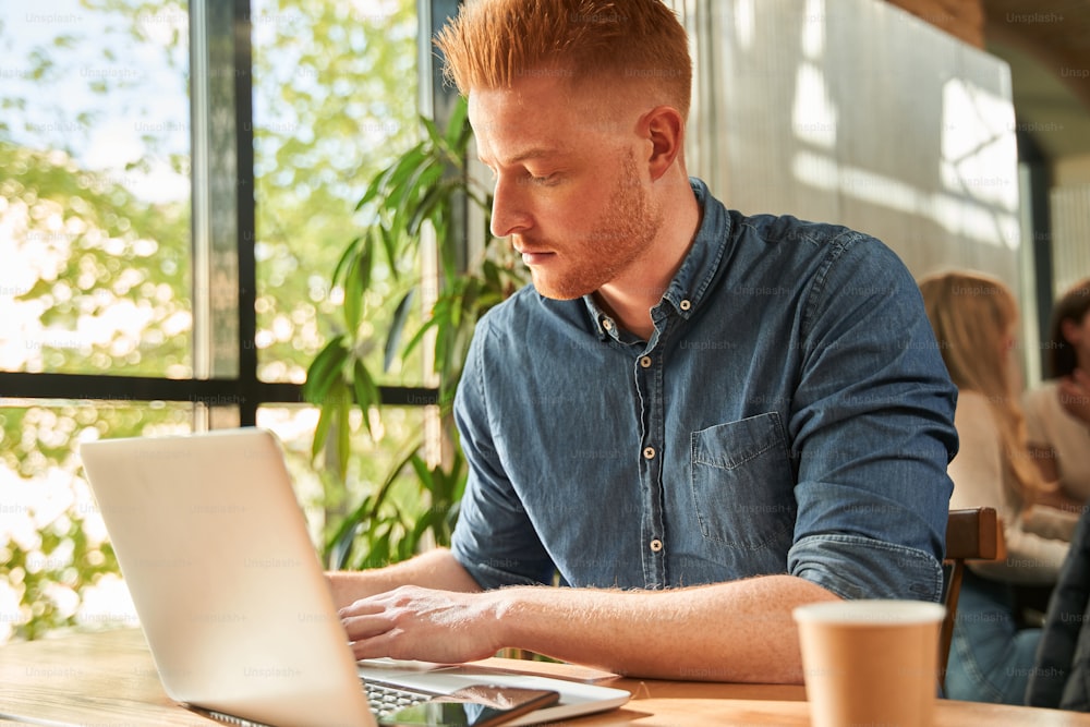 Jovem elegante vestindo camisa jeans sendo focado enquanto sentado no café e trabalhando em laptop pela manhã. Conceito de freelancers de pessoas