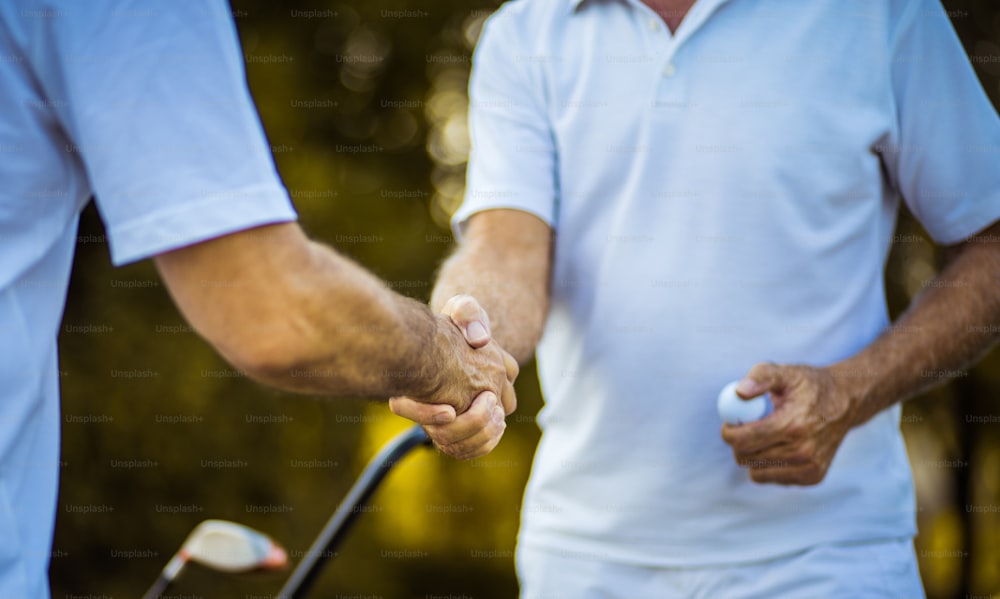 Two older men stand on a golf course and talk. Focus is on hands.
