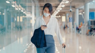 アジアのビジネスガールは、空港の国内線でターミナルまで荷物を持って搭乗券を歩くチェックインにスマートフォンを使用します。ビジネス通勤者のcovidパンデミック、ビジネス旅行の社会的距離のコンセプト。
