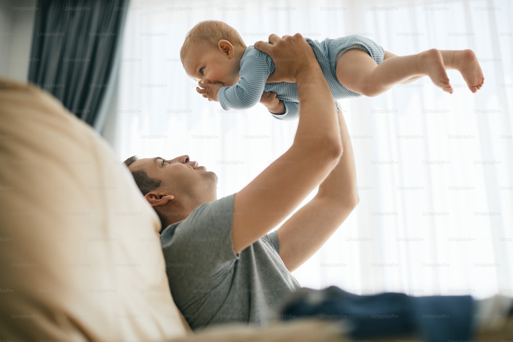 Young father holding his baby son high up while sitting on the sofa and having fun at home.