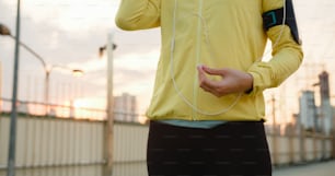 Beautiful young Asia athlete lady exercises using smartphone for listen to music while running in urban environment. Korean teen girl wearing sports clothes on walkway bridge in early morning.