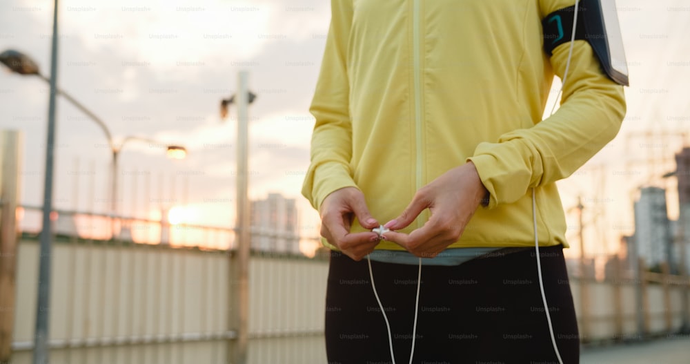 美しい若いアジアのアスリートの女性は、都市環境で走りながら音楽を聴くためにスマートフォンを使用してエクササイズします。早朝に歩道の橋でスポーツ服を着ている韓国の十代の女の子。