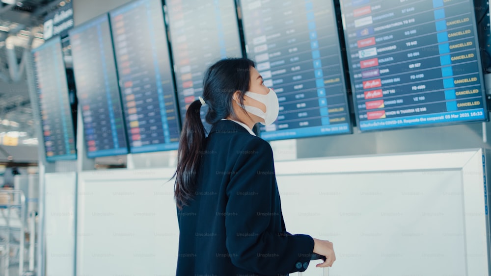 아시아 비즈니스 소녀는 국제 공항에서 그녀의 비행을 확인하는 정보를 보는 보드 앞에 여행 가방 스탠드와 얼굴 마스크를 착용. 비즈니스 통근자 covid 전염병, 비즈니스 여행 개념.