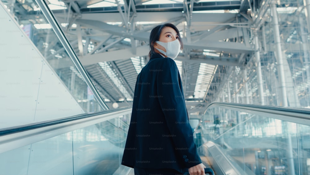 Asian business girl wear face mask drag luggage stand on escalator look around walk to terminal at international airport. Business commuter covid pandemic, Business travel social distancing concept.