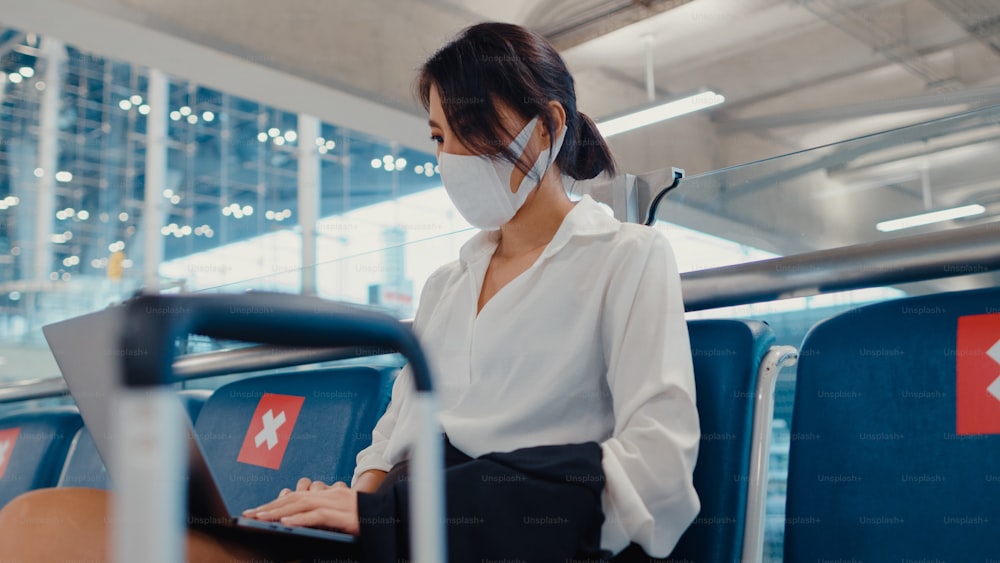 Asian business lady traveller wear face mask sitting in bench use laptop for work between wait for flight in terminal at airport. Business travel commuter in covid pandemic, Business travel concept.
